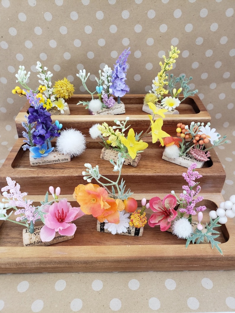 May include: Assortment of small floral arrangements on cork rounds, displayed on a tiered wooden stand. The arrangements feature a variety of flowers, including purple, yellow, pink, and orange.