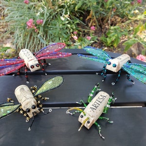 Puede incluir: Cuatro insectos decorativos hechos de corchos de vino y cuentas. Los insectos tienen alas de alambre y cuentas en tonos rojo, azul, verde y dorado. Los corchos est&aacute;n etiquetados con las palabras "Mo&euml;t" y "Artesa".