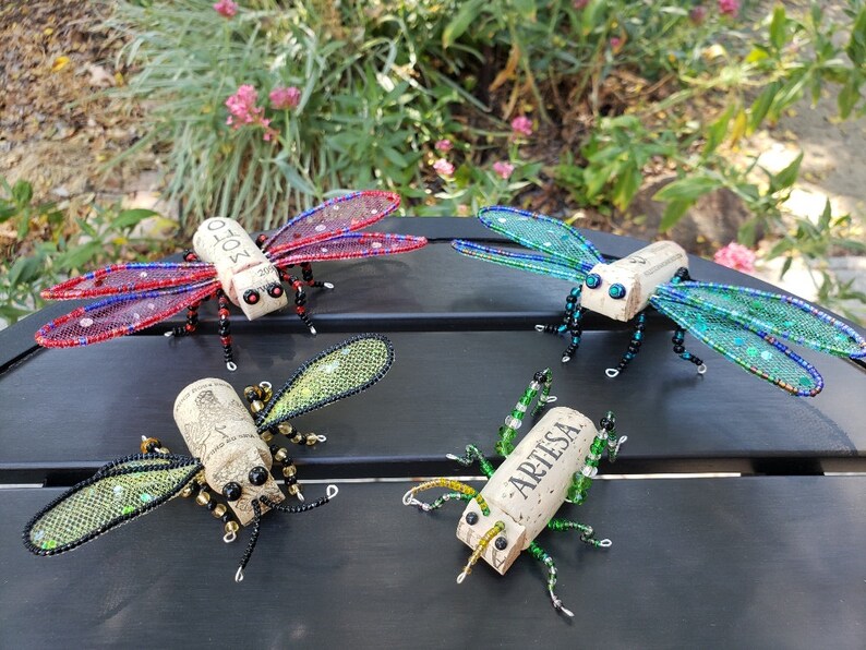 Puede incluir: Cuatro insectos decorativos hechos de corchos de vino y cuentas. Los insectos tienen alas de alambre y cuentas en tonos rojo, azul, verde y dorado. Los corchos est&aacute;n etiquetados con las palabras "Mo&euml;t" y "Artesa".