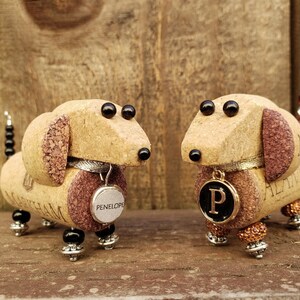 May include: Two miniature dachshund figurines made from wine corks. The figurines are brown and have black eyes. One figurine has a tag that says "PENELOPE" and the other has a tag with the letter "P".