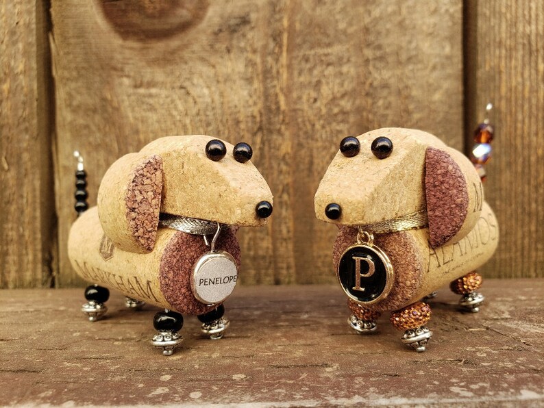 May include: Two miniature dachshund figurines made from wine corks. The figurines are brown and have black eyes. One figurine has a tag that says "PENELOPE" and the other has a tag with the letter "P".