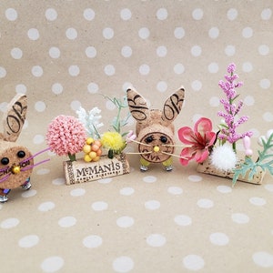 May include: Three cork bunny figurines with pink and white flowers, a pink flower, and white berries. The bunnies have black eyes and a yellow nose. The cork is from a wine bottle and has the text "McManis Family Vineyards" on it.