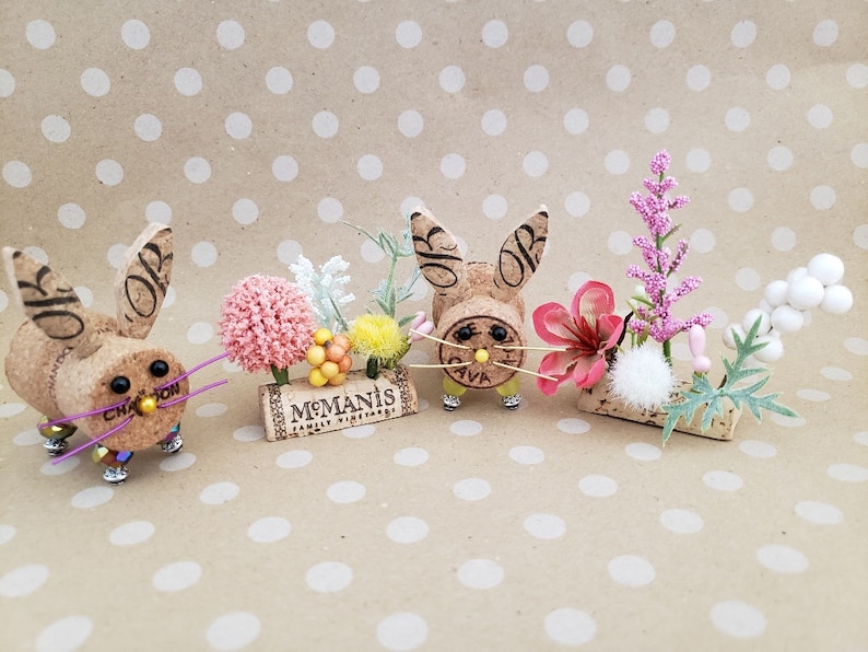 May include: Three cork bunny figurines with pink and white flowers, a pink flower, and white berries. The bunnies have black eyes and a yellow nose. The cork is from a wine bottle and has the text "McManis Family Vineyards" on it.