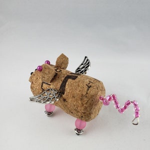 May include: A small, winged pig figurine made from a wine cork. The pig has pink beads for eyes and feet, and a pink beaded tail. The wings are made of silver metal.