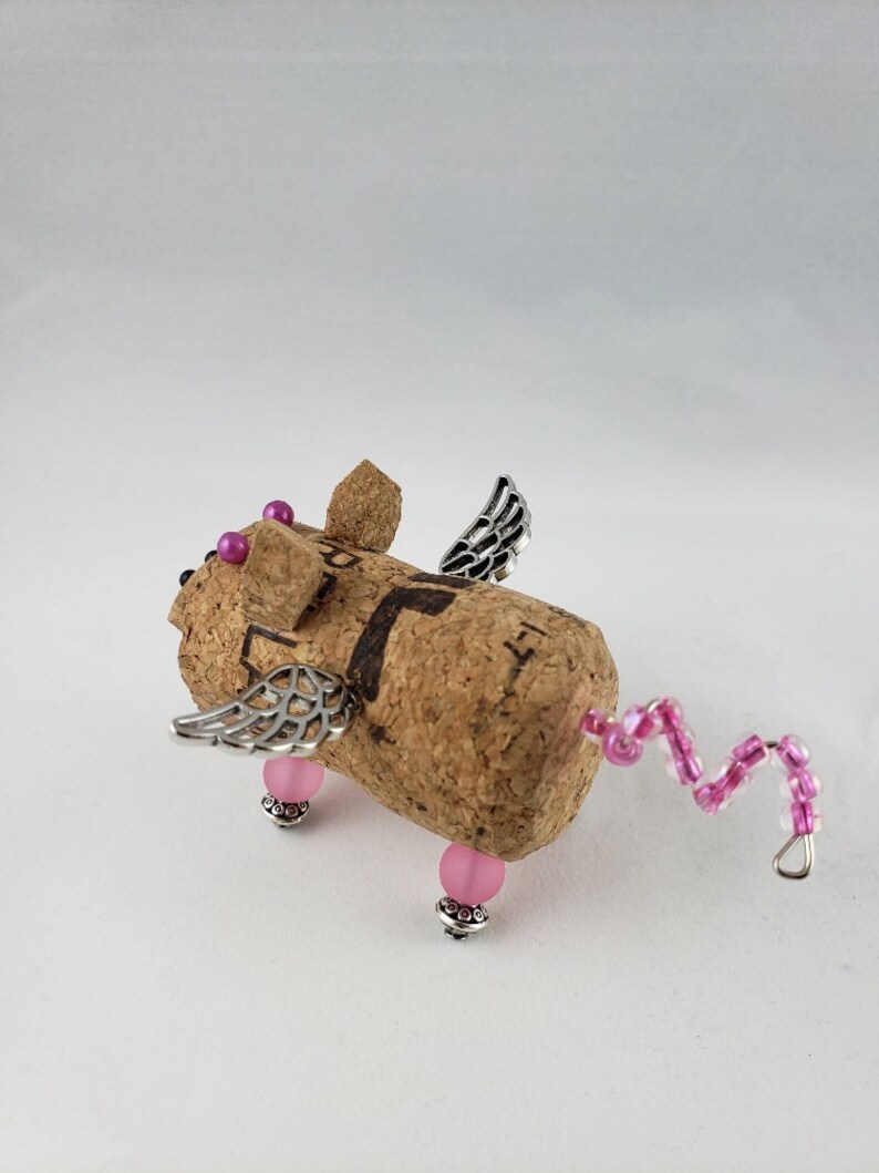 May include: A small, winged pig figurine made from a wine cork. The pig has pink beads for eyes and feet, and a pink beaded tail. The wings are made of silver metal.