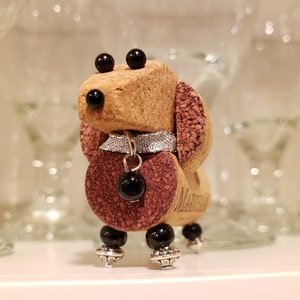 May include: A small, handcrafted dachshund figurine made from wine corks. The figurine is decorated with black beads for eyes, a silver collar with a black bead charm, and black beads for feet. The dachshund is standing on a white surface.