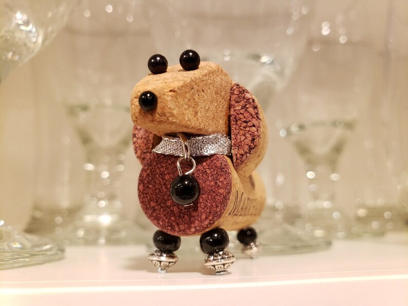 May include: A small, handcrafted dachshund figurine made from wine corks. The figurine is decorated with black beads for eyes, a silver collar with a black bead charm, and black beads for feet. The dachshund is standing on a white surface.