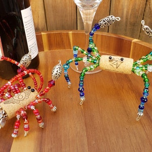 May include: Two beaded wine bottle stoppers shaped like crabs. One is red and the other is green and blue. Both have cork bodies and silver accents.