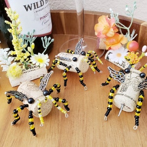 Puede incluir: Cuatro tapones de botella de abeja hechos a mano de corcho y cuentas negras y amarillas. Cada abeja tiene alas plateadas y ojos de cuentas negras. Las abejas est&aacute;n sentadas sobre una superficie de madera.