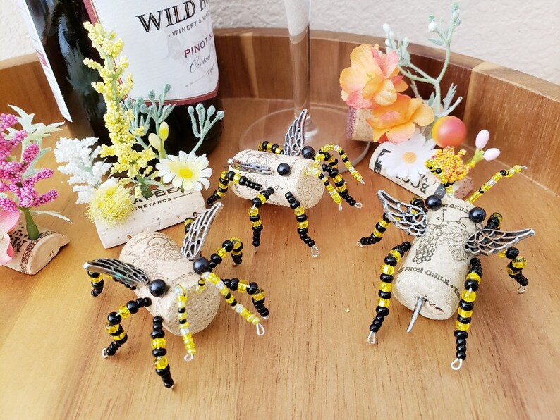 Puede incluir: Cuatro tapones de botella de abeja hechos a mano de corcho y cuentas negras y amarillas. Cada abeja tiene alas plateadas y ojos de cuentas negras. Las abejas est&aacute;n sentadas sobre una superficie de madera.