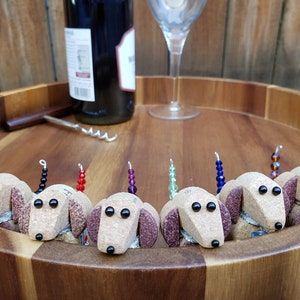 May include: Eight wine bottle stoppers shaped like brown and white dachshunds with black eyes. Each stopper has a different colored beaded tail.