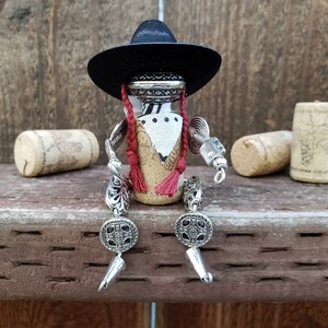 May include: A whimsical figurine made from a wine cork, dressed in a black hat, white fabric, and red tassels. The figurine has silver metal arms and legs, and is sitting on a wooden surface.