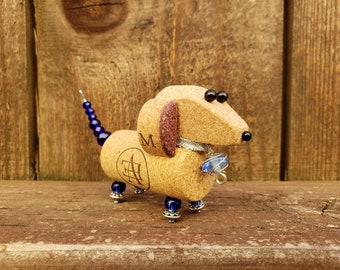May include: A small dachshund figurine made from wine corks, with blue beads for a tail and legs, and a blue glass charm around its neck. The figurine has black eyes and a brown nose.