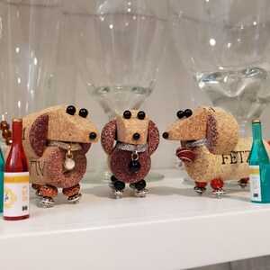 May include: Three miniature dachshund figurines made from wine corks, each with a different colored collar and a small bottle of wine. The figurines are standing on a white surface.