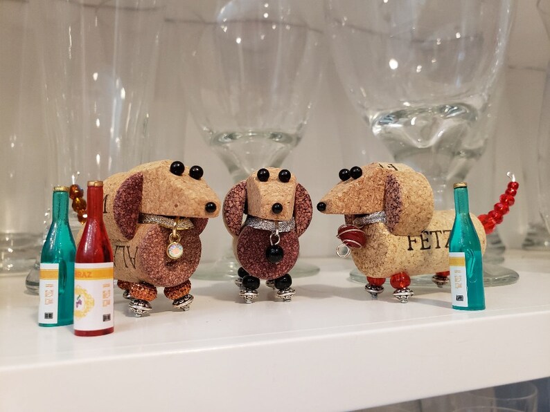 May include: Three miniature dachshund figurines made from wine corks, each with a different colored collar and a small bottle of wine. The figurines are standing on a white surface.