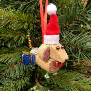 May include: A cork dachshund Christmas ornament wearing a red Santa hat with white trim and holding a blue gift box. The ornament is decorated with gold glitter and red and green beads.