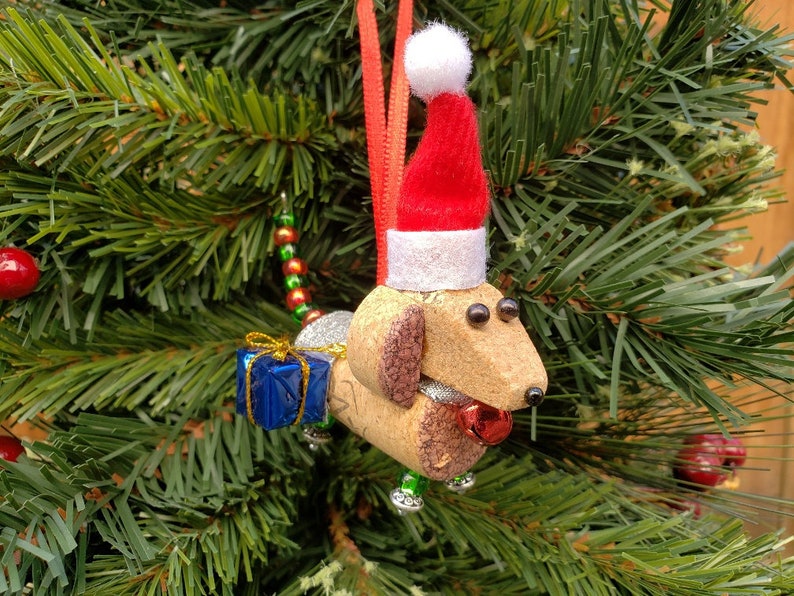May include: A cork dachshund Christmas ornament wearing a red Santa hat with white trim and holding a blue gift box. The ornament is decorated with gold glitter and red and green beads.
