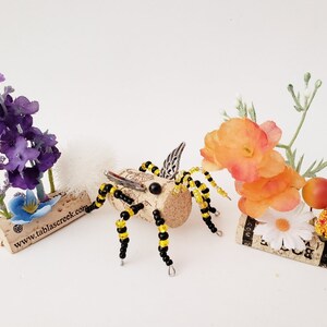 Puede incluir: Una abeja de cuentas con cuentas negras y amarillas y alas plateadas se sienta sobre un corcho con el texto "www.tablascreek.com". Otros dos corchos est&aacute;n decorados con flores artificiales, incluidas flores moradas, naranjas y blancas.