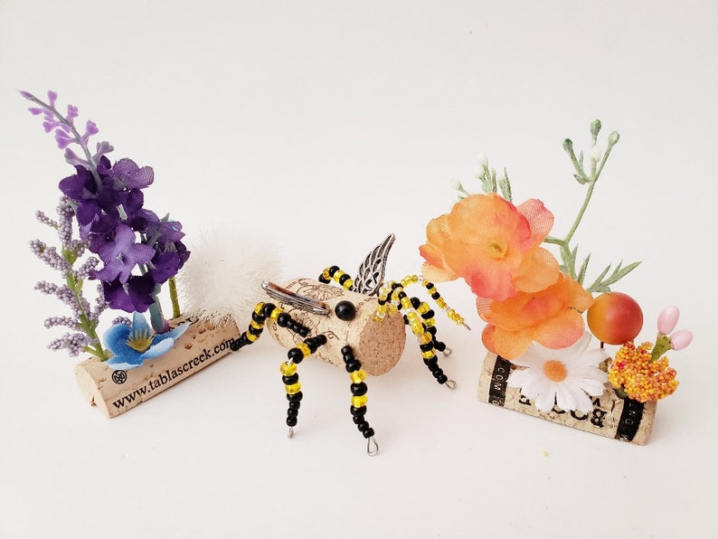 Puede incluir: Una abeja de cuentas con cuentas negras y amarillas y alas plateadas se sienta sobre un corcho con el texto "www.tablascreek.com". Otros dos corchos est&aacute;n decorados con flores artificiales, incluidas flores moradas, naranjas y blancas.