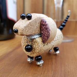 May include: A small dachshund dog figurine made from a wine cork with black beads for eyes and legs. The dog has a silver collar with a black bead charm and a pink cork ear.