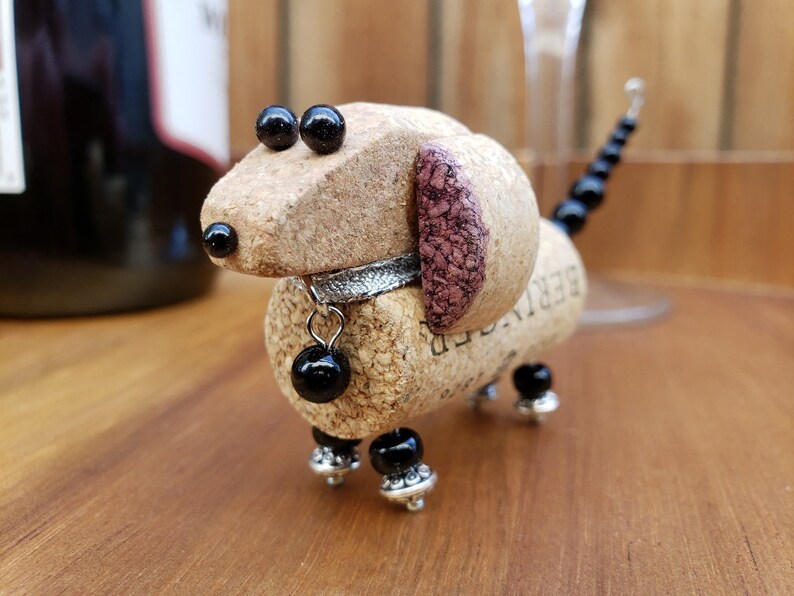 May include: A small dachshund dog figurine made from a wine cork with black beads for eyes and legs. The dog has a silver collar with a black bead charm and a pink cork ear.