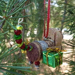 May include: A Christmas ornament made from wine corks, shaped like a dog carrying a small green gift box. The dog has a red and white striped ribbon attached to its head. The ornament is decorated with red, green, and gold beads.