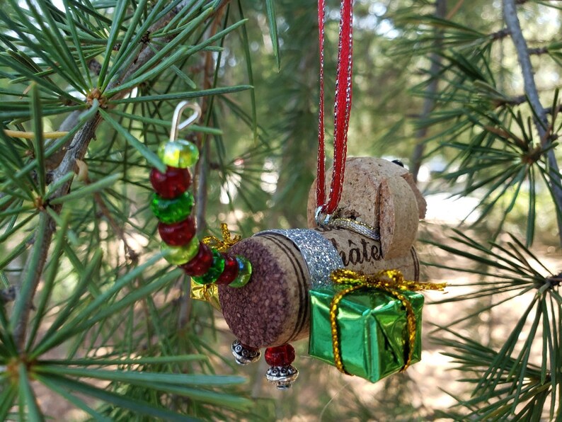 May include: A Christmas ornament made from wine corks, shaped like a dog carrying a small green gift box. The dog has a red and white striped ribbon attached to its head. The ornament is decorated with red, green, and gold beads.