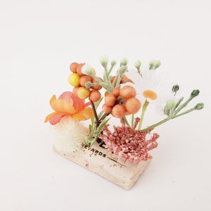 May include: A small floral arrangement with white, orange, and yellow flowers, and orange berries, in a cork container with the word "Rewards" printed on it.
