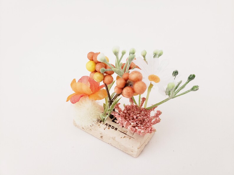 May include: A small floral arrangement with white, orange, and yellow flowers, and orange berries, in a cork container with the word "Rewards" printed on it.