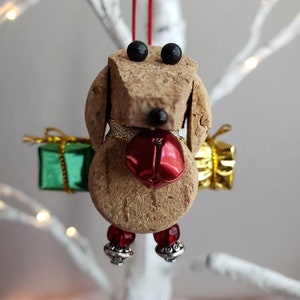 May include: A cork dog ornament with black eyes, a red bell, and green and gold gift boxes. The dog is wearing red beads and silver bells on its feet.
