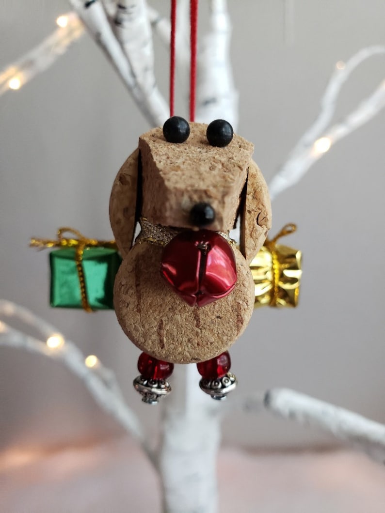 May include: A cork dog ornament with black eyes, a red bell, and green and gold gift boxes. The dog is wearing red beads and silver bells on its feet.