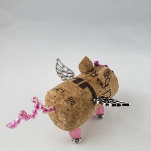 May include: A small, winged pig figurine made from a wine cork. The pig has pink beads for legs and a pink beaded tail. The wings are silver and the pig has pink beads for eyes.