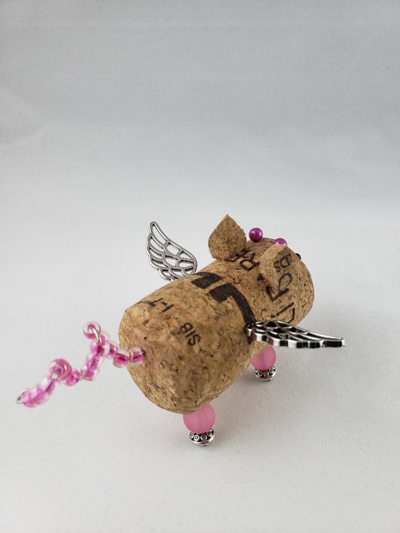 May include: A small, winged pig figurine made from a wine cork. The pig has pink beads for legs and a pink beaded tail. The wings are silver and the pig has pink beads for eyes.