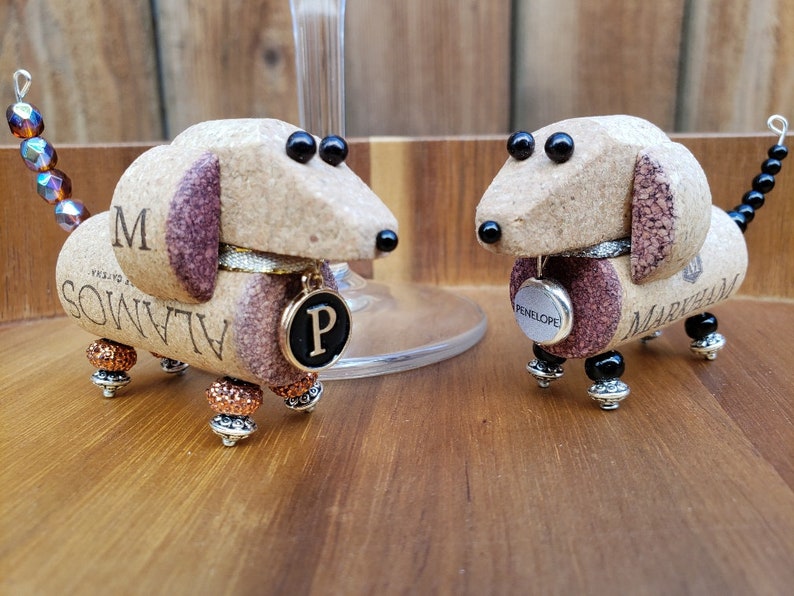 May include: Two dachshund-shaped ornaments made from wine corks. The corks are painted brown and have black eyes and noses. The ornaments have beaded tails and are decorated with beads and charms. One ornament has a charm that says "P" and the other has a charm that says "Penelope".
