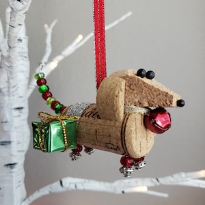 May include: A cork dachshund Christmas ornament with a green gift box, red beads, and a red bell. The ornament is hanging from a red ribbon.