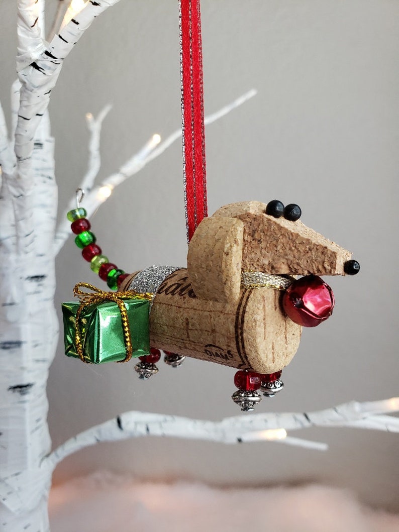 May include: A cork dachshund Christmas ornament with a green gift box, red beads, and a red bell. The ornament is hanging from a red ribbon.