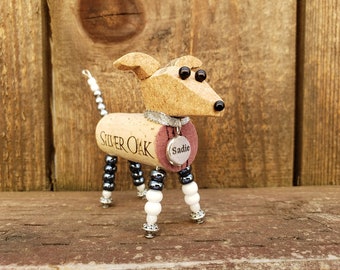 May include: A small, handcrafted dog figurine made from a wine cork. The dog has black and white beads for legs and a silver collar with a tag that reads "Sadie". The cork is labeled "Silver Oak".
