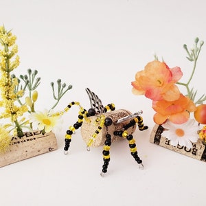 Puede incluir: Una abeja de cuentas negras y amarillas con alas de metal se sienta sobre un corcho con el texto "AT BER" y "VINEYARDS". Otros dos corchos est&aacute;n decorados con peque&ntilde;as flores amarillas y naranjas.