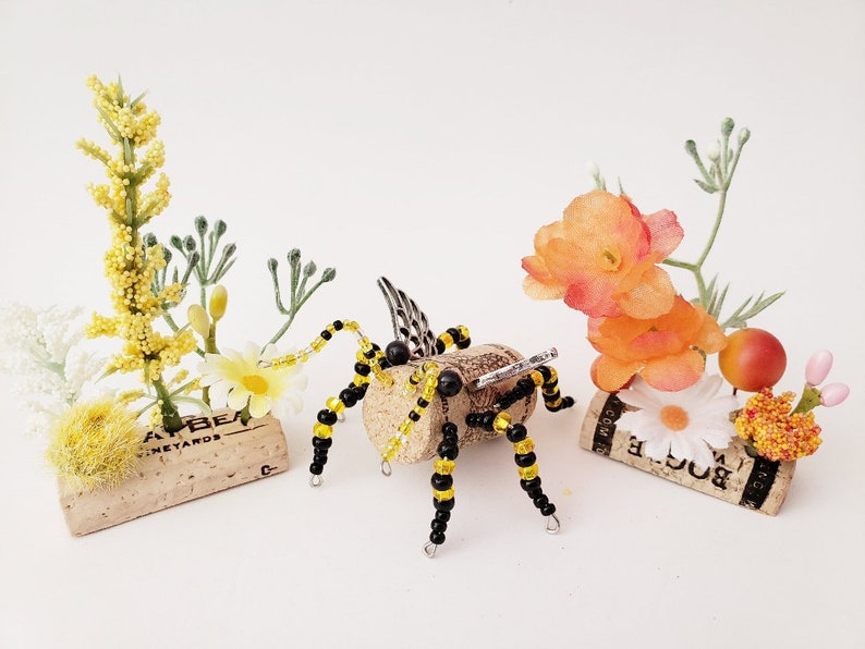 Puede incluir: Una abeja de cuentas negras y amarillas con alas de metal se sienta sobre un corcho con el texto "AT BER" y "VINEYARDS". Otros dos corchos est&aacute;n decorados con peque&ntilde;as flores amarillas y naranjas.