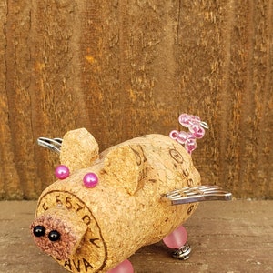 May include: A whimsical pig figurine made from a wine cork, with pink and black beads for eyes and legs, and silver wings. The cork has the text "CEST'A" and "AVA" printed on it.