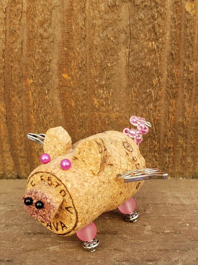 May include: A whimsical pig figurine made from a wine cork, with pink and black beads for eyes and legs, and silver wings. The cork has the text "CEST'A" and "AVA" printed on it.