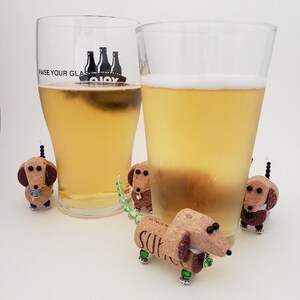 May include: Four miniature dachshund figurines made from wine corks, with green and blue beaded tails, are arranged around two glasses of beer. One glass has the text "Raise Your Glass" printed on it.
