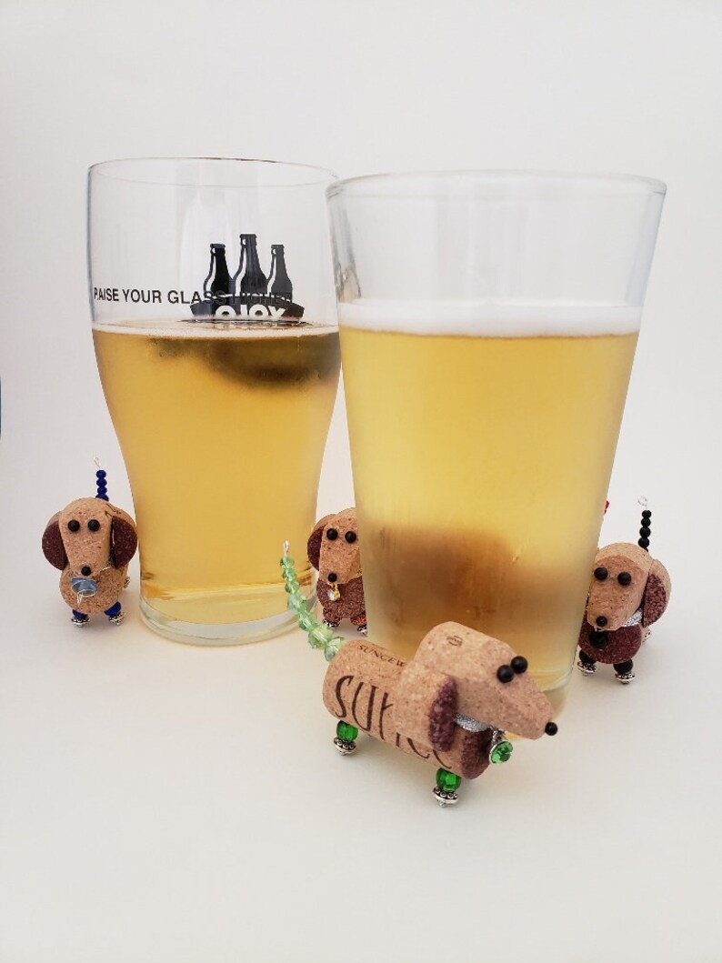 May include: Four miniature dachshund figurines made from wine corks, with green and blue beaded tails, are arranged around two glasses of beer. One glass has the text "Raise Your Glass" printed on it.