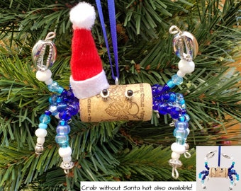 May include: A blue and white beaded crab ornament with a red Santa hat on top of a cork. The ornament is hanging from a blue ribbon. "Crab without Santa hat also available!" is written below the ornament.