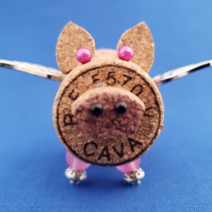 May include: A cork pig with pink ears and wings. The pig is made from a wine cork with the words "BEST 70" and "CAVA" printed on it. The pig has pink beads for legs and silver wings.