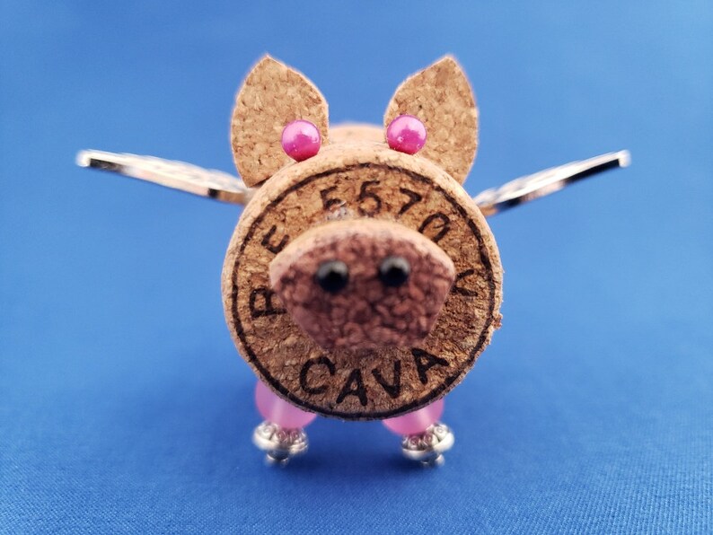 May include: A cork pig with pink ears and wings. The pig is made from a wine cork with the words "BEST 70" and "CAVA" printed on it. The pig has pink beads for legs and silver wings.