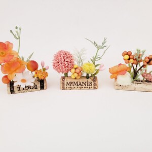 May include: Three small floral arrangements in cork containers. The first arrangement features orange and white flowers with green stems. The second arrangement features pink, yellow, and white flowers with green stems. The third arrangement features orange and white flowers with brown stems and berries.