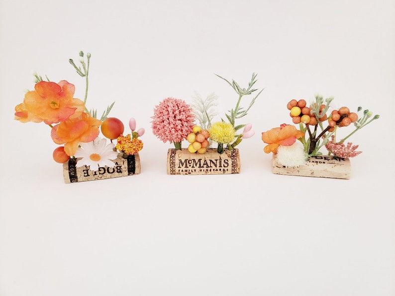 May include: Three small floral arrangements in cork containers. The first arrangement features orange and white flowers with green stems. The second arrangement features pink, yellow, and white flowers with green stems. The third arrangement features orange and white flowers with brown stems and berries.