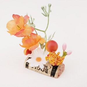 May include: A small floral arrangement in a cork with orange, white, and pink flowers. The cork has the word "BOGLI" printed on it.