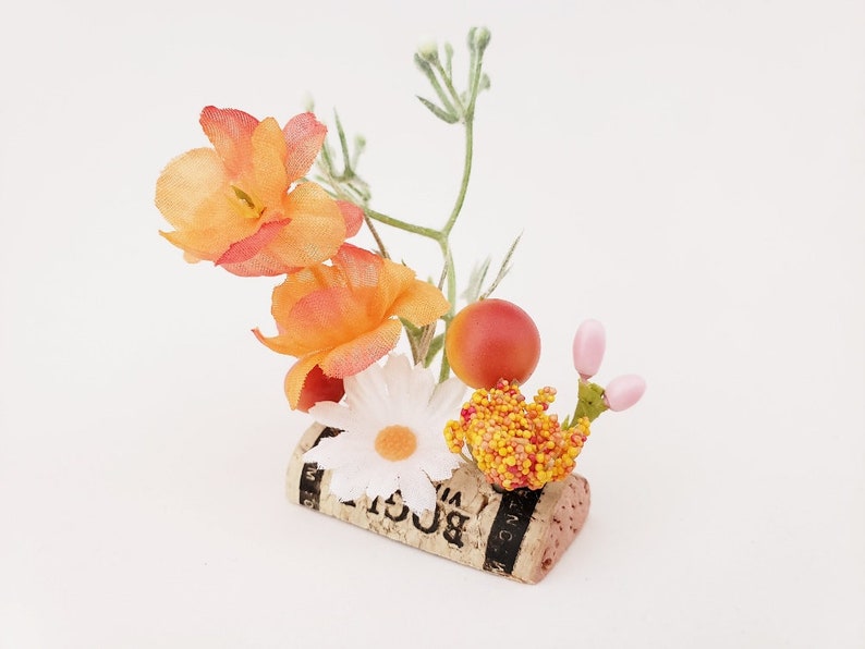 May include: A small floral arrangement in a cork with orange, white, and pink flowers. The cork has the word "BOGLI" printed on it.